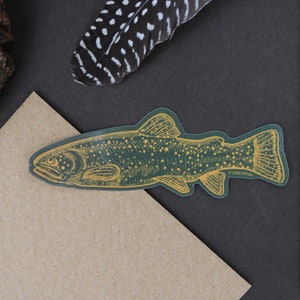 May include: A green and gold fish sticker with the text "A Little Wilder". The sticker is placed on a brown envelope, with a piece of wood and a feather in the background. The sticker is a detailed illustration of a fish.