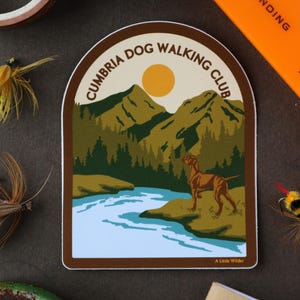 May include: A die-cut sticker featuring a brown dog standing by a blue river with green mountains and a yellow sun in the background. The text CUMBRIA DOG WALKING CLUB is arched at the top, and the text A Little Wilder is printed at the bottom.
