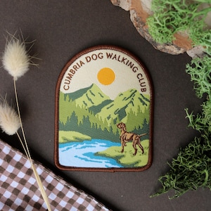May include: A brown embroidered patch with a white background. The patch features a mountain scene with a river, a sun, and a brown dog. The text "Cumbria Dog Walking Club" is written in a brown circle around the scene.