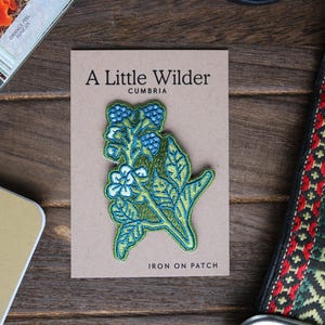 May include: A green and blue embroidered iron-on patch featuring a stylised flower with the text "A Little Wilder Cumbria Iron On Patch".