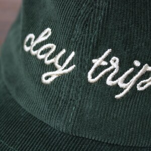 May include: A dark green corduroy baseball cap with white embroidered text that reads "day trip".