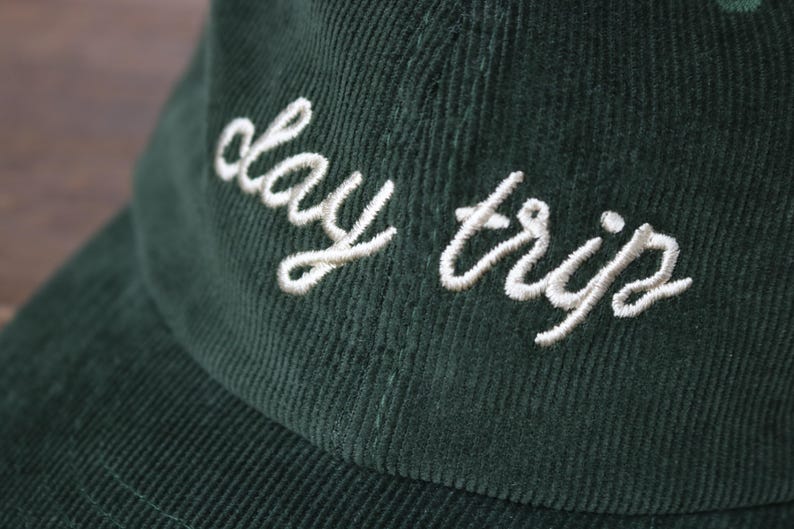 May include: A dark green corduroy baseball cap with white embroidered text that reads "day trip".