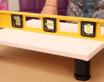May include: A white rectangular canvas on black legs is being levelled with a yellow spirit level. The level has three bubble vials. Paint tubes are visible in the background.