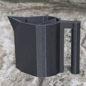 Puede incluir: Una jarra gris oscura impresa en 3D con asa. La jarra tiene un diseño geométrico único con un pico puntiagudo.