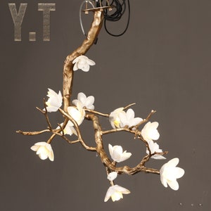 May include: A gold-colored, branch-shaped chandelier with white flower-shaped lights. The fixture has a natural, organic design, with the lights resembling blossoms. The letters "Y.T" are visible in the upper left corner.