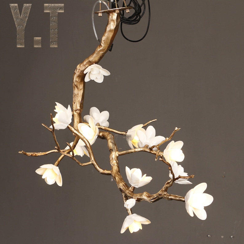 May include: A gold-colored, branch-shaped chandelier with white flower-shaped lights. The fixture has a natural, organic design, with the lights resembling blossoms. The letters "Y.T" are visible in the upper left corner.