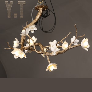 May include: A decorative chandelier in a branch design with white flower-shaped lights. The fixture has a gold-colored finish and includes the letters "Y.T" in the upper left corner.