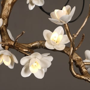 May include: Close-up of a decorative chandelier featuring white porcelain flower blossoms with warm-toned light bulbs. The fixture has gold-colored branches and a dark-colored support structure, creating an elegant floral design.