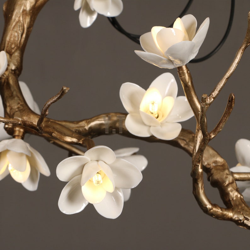 May include: Close-up of a decorative chandelier featuring white porcelain flower blossoms with warm-toned light bulbs. The fixture has gold-colored branches and a dark-colored support structure, creating an elegant floral design.