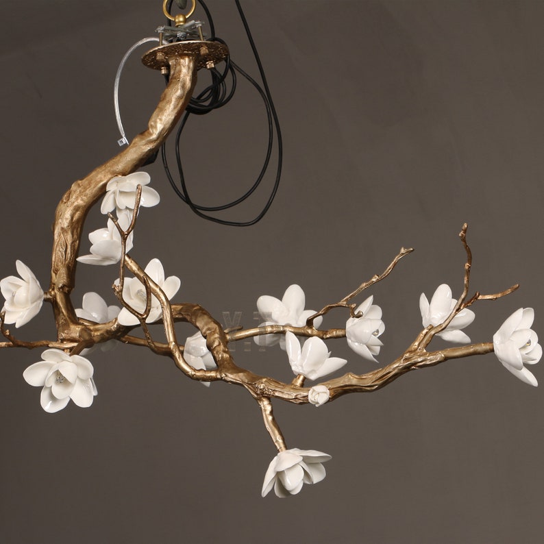 May include: A gold-colored, branch-shaped chandelier with white flower accents. The fixture has a central mounting point with wires and a clear plastic support. The flowers are clustered along the branches, creating a natural, elegant design.