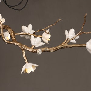 May include: A decorative chandelier featuring a gold-colored branch design with white flower-shaped lights. The fixture has a natural, organic aesthetic, with the flowers emitting a soft, warm glow. The overall design is elegant and artistic.