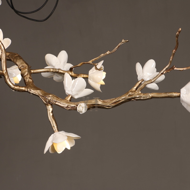 May include: A decorative chandelier featuring a gold-colored branch design with white flower-shaped lights. The fixture has a natural, organic aesthetic, with the flowers emitting a soft, warm glow. The overall design is elegant and artistic.