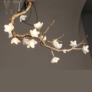 May include: A gold-toned, branch-shaped chandelier with white, illuminated flower-shaped lights. The fixture is suspended from the ceiling, with the text "Y.T." visible in the upper left corner. The design evokes a natural, floral theme.