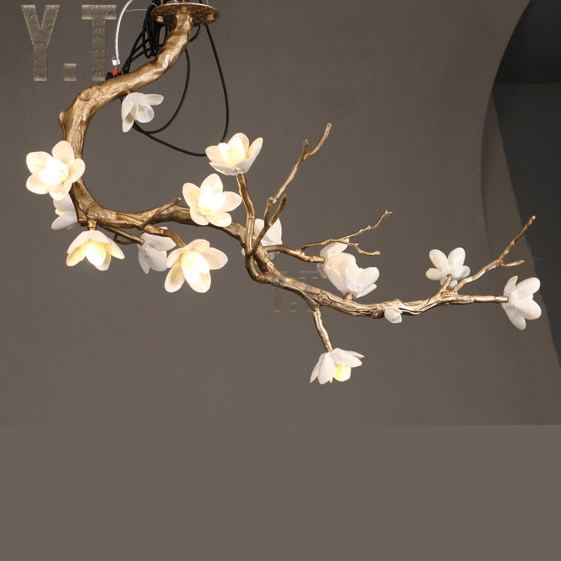 May include: A gold-toned, branch-shaped chandelier with white, illuminated flower-shaped lights. The fixture is suspended from the ceiling, with the text "Y.T." visible in the upper left corner. The design evokes a natural, floral theme.