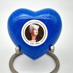 May include: Blue heart-shaped cremation urn with a silver stand. A photo of a woman is in a circular window on the urn. The text "Sally Davies 1939 - 2016" is printed below the photo.
