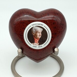 May include: A red heart-shaped urn with a white circle containing a photo of a woman and the text "SALLY DAVIES 1939 - 2016". The urn is on a silver stand.