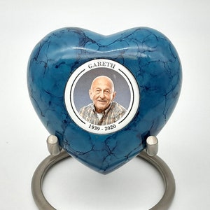May include: A blue heart-shaped cremation urn with a silver stand. The urn has a white circle with a photo of a man and the text "Gareth 1939 - 2020".