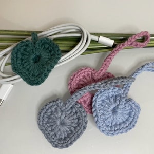 Könnte beinhalten: Gehäkelte Kabelorganisatoren in Herzform und ein Blatt. Die Organizer sind in Grün-, Rosa-, Blau- und Grautönen gehalten. Ein weißes Kabel mit USB-C-Anschluss ist ebenfalls vorhanden. Die Artikel liegen auf einer weißen Oberfläche.