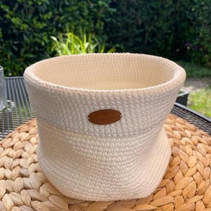 Könnte beinhalten: Cremefarbener, handgestrickter Korb mit breiter, offener Oberseite. Der Korb hat eine zylindrische Form mit leicht ausgestelltem Boden. Ein kleines, ovales braunes Lederetikett ist vorne angebracht. Der Korb steht auf einer gewebten, runden Matte.
