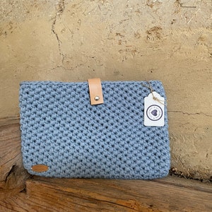 Könnte beinhalten: Eine hellblaue gehäkelte Clutch mit Lederriemenverschluss und kleinem Anhänger. Die handgefertigte Tasche ist rechteckig und hat ein strukturiertes Stichmuster. Die Clutch liegt auf einer Holzoberfläche.