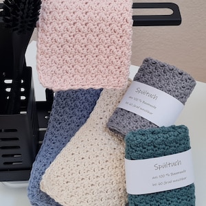 Könnte beinhalten: Vier gehäkelte Geschirrtücher in verschiedenen Farben: rosa, beige, blau und grün. Jedes Geschirrtuch hat ein Etikett mit der Aufschrift "Spültuch aus 100% Baumwolle bei 60 Grad waschbar".