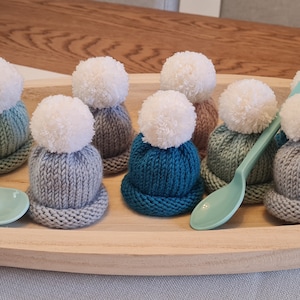 Könnte beinhalten: Eine Sammlung von 12 gestrickten Eierwärmern mit Bommeln in verschiedenen Farben, darunter Türkis, Grau und Rosa. Die Eierwärmer sind auf einem Holztablett mit zwei blauen Löffeln angeordnet.