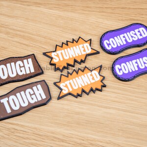 Puede incluir: Seis parches de fieltro con diferentes palabras. Dos parches marrones dicen "TOUGH", dos parches naranjas dicen "STUNNED" y dos parches morados dicen "CONFUSED".