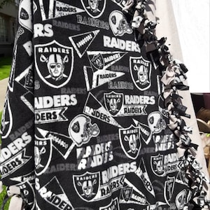 May include: A black and white fleece tie blanket with a repeating pattern of the Las Vegas Raiders logo. The blanket has a fringe border.