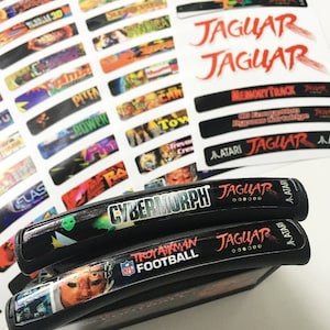 May include: Two black Atari Jaguar game cartridges with labels. The top cartridge has a black and green label with the text "CYBERMORPH" in white. The bottom cartridge has a black label with a football player on it and the text "TROY AIKMAN FOOTBALL" in white.