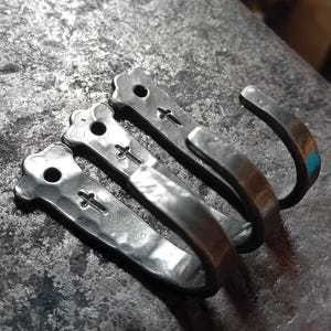May include: Four handcrafted metal hooks with a hammered texture. Each hook features a cross design and a small hole. The fourth hook has a turquoise accent. The hooks are arranged on a textured metal surface.