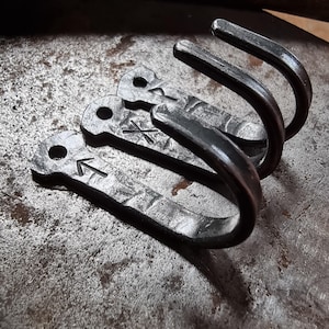 Peut inclure: Trois crochets en métal forgé à la main, d'esthétique rustique et industrielle. Chaque crochet a un design incurvé et un dessus plat avec un trou pour le montage. Le métal a une finition sombre et texturée, et certains crochets présentent des symboles gravés.