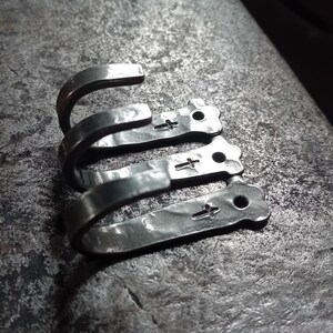 May include: Close-up of three silver metal hooks with a hammered texture. Each hook has a cross-shaped design and a small hole. The hooks are stacked on top of each other, with the curved ends facing upwards.