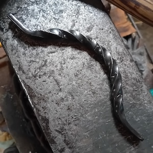 Peut inclure: Une tige métallique torsadée, probablement forgée à la main, est posée sur une surface métallique texturée. La tige est gris foncé et présente une finition lisse et légèrement brillante.