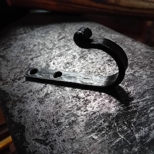 Può includere: Un gancio in metallo nero con un decoro a forma di rotolo nella parte superiore. Il gancio è montato su una superficie piana.