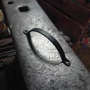 以下が含まれることがあります： 手作りの黒い金属製のハンドルで、曲線的なデザインです。 ハンドルには取り付け用の小さな穴が2つあり、おそらく金床である、質感のある灰色の金属表面に置かれています。 ハンドルの形状から、引き出しやキャビネットに使用できることが示唆されています。