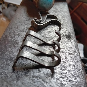 May include: Close-up of several small, silver-coloured metal clasps, possibly for jewellery or crafts. The clasps have a curved design with a hook shape. The metal has a slightly aged appearance, suggesting a vintage or handmade quality. The clasps are arranged on a textured, grey metal surface.