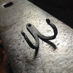 May include: Hand-forged, dark grey metal hook with two curved prongs and a single hole for mounting. The hook is made of iron and sits on a textured metal surface, showcasing the craftsmanship of the piece.