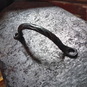 May include: A hand-forged, black metal handle with a curved design. The handle features a textured surface and is attached to a flat base with two mounting holes. The metal has a dark, aged appearance, suggesting a rustic or industrial aesthetic.