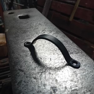 Könnte beinhalten: Ein schwarzer, gebogener Metallgriff mit zwei Befestigungslöchern. Der Griff ist auf einer strukturierten, grauen Metalloberfläche, wahrscheinlich einem Amboss, abgebildet. Das Design des Griffs deutet darauf hin, dass er für einen Schubladengriff oder eine ähnliche Hardware verwendet werden könnte.