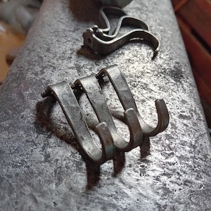 May include: Three metal hooks with a curved design, arranged in a row. The hooks are a dull silver colour and appear to be handmade. A decorative metal piece is in the background. The items are on a textured, grey surface.