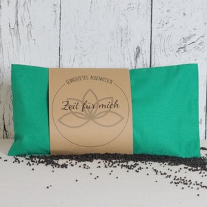 Könnte beinhalten: Ein rechteckiges, türkisfarbenes Augenpolster mit einem braunen Etikett mit dem Text "Zeit für mich" und einem floralen Design. Das Kissen ist von kleinen, dunklen Samen umgeben, wahrscheinlich zur Gewichtung.