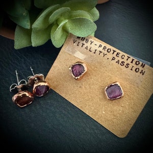 May include: A pair of earrings with small, square-shaped, copper settings and pink, faceted gemstones. The earrings are displayed on a brown card with the text "Ruby - Protection, Vitality, Passion".