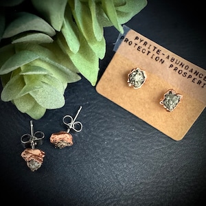 May include: A pair of gold-plated earrings with raw pyrite stones. The earrings are on a brown card with the text "Pyrite - Abundance, Protection, Prosperity".