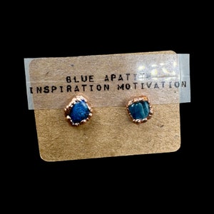 May include: A pair of blue apatite earrings with gold settings. The earrings are on a brown card with the text "BLUE APATITE INSPIRATION MOTIVATION".