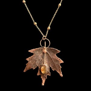 May include: A copper-toned maple leaf pendant hangs from a delicate gold-colored chain with small bead accents. The leaf features a textured surface and a single, small, translucent amber-colored stone embedded near the center of the leaf base.