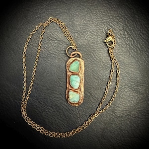 May include: A handcrafted copper pendant necklace featuring three turquoise-colored stones. The pendant is rectangular with a textured surface and is suspended from a delicate gold-colored chain. The necklace is displayed against a dark background.