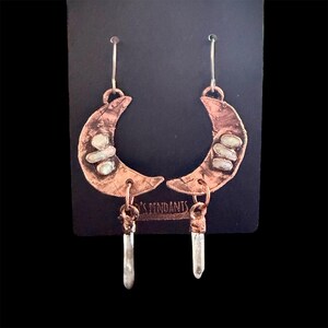 May include: Copper crescent moon earrings with white stone accents and crystal pendants. The earrings hang from silver hooks and are displayed on a black card. The earrings are handmade.