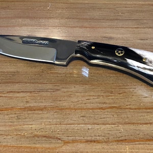 Op de afbeelding: Een zilveren mes met een zwart-witte handvat. Het handvat heeft gouden accenten en een kleine gouden cirkel met een ontwerp in het midden.