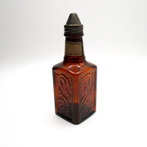 Puede incluir: Una botella de vidrio marr&oacute;n con un tap&oacute;n de metal y un dise&ntilde;o floral en relieve. La botella tiene una etiqueta en la parte delantera.