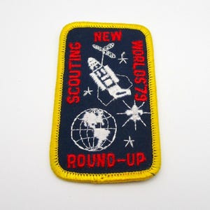 May include: A rectangular patch with a navy blue background and a yellow border. The patch features the words "SCOUTING" and "ROUND-UP" in red, and "NEW WORLDS '79" in red. A white rocket, globe, and stars are also present.